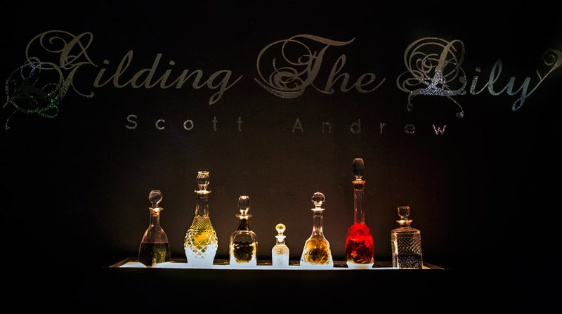 Seven ornate glass bottles with colorful liquids, illuminated on a dark shelf beneath "Gilding The Lily Scott Andrew" in scri