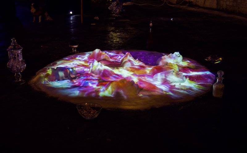 Installation with sugar mound illuminated by swirling pink, purple, and yellow lights, with glass vessels nearby.