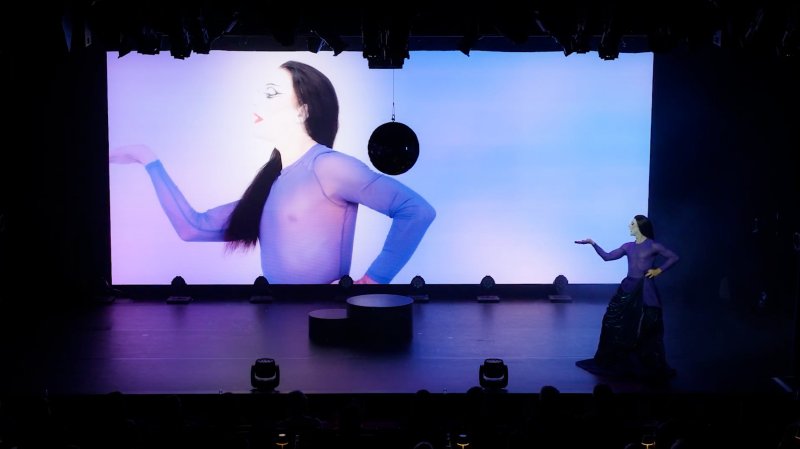 Performer in purple sheer top and dark skirt poses on stage, mirroring a large screen projection of themselves.