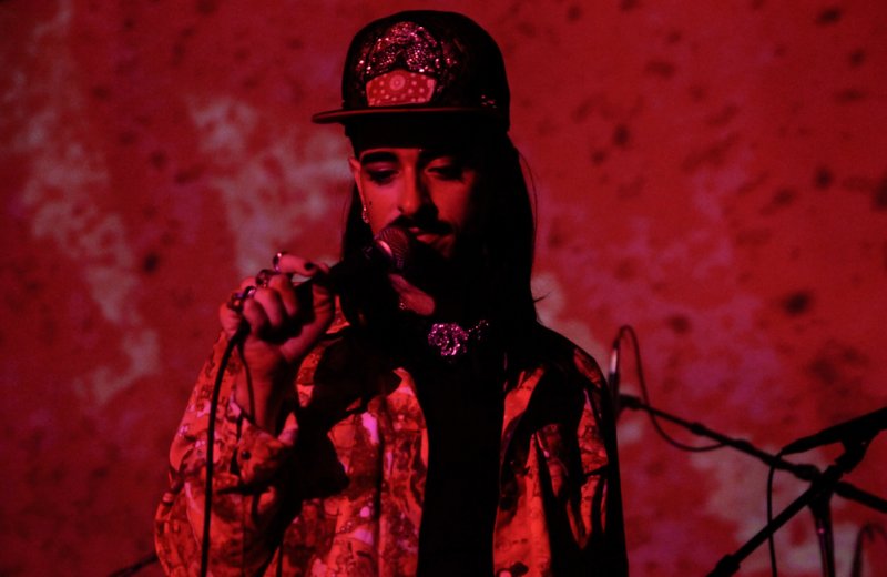 Performer in a sequined cap and patterned jacket sings into a microphone, bathed in dramatic red light against a textured red
