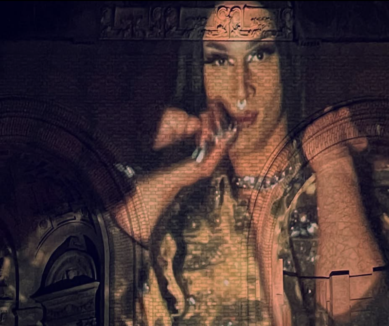 Drag queen with dark hair and makeup, nose ring, and ornate top, projected onto a brick wall with arched doorways and carved details.