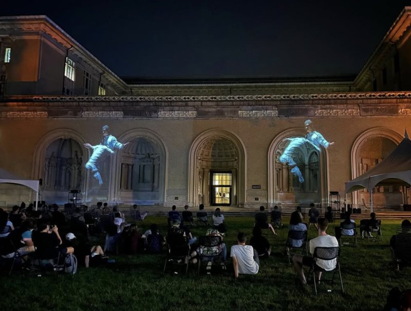 Dragon Sisters" by Scott N Andrew. Two pale blue digital avatars projected onto a building's facade at night, viewed by an audience on a lawn.