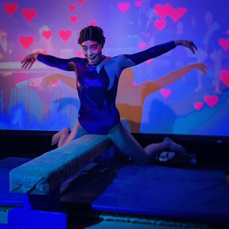 A person in a blue leotard balances on a low beam, arms outstretched, against a wall projected with pink hearts.