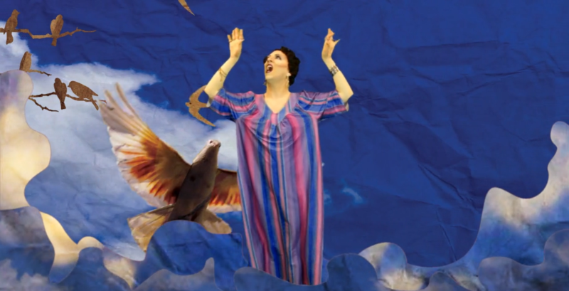 Drag queen in striped caftan with arms raised, surrounded by paper cut-out birds and clouds against a blue sky.