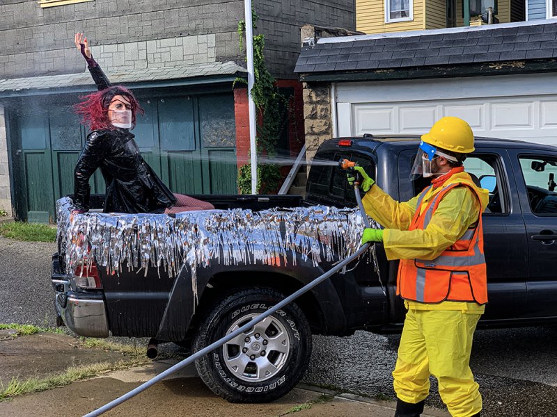 A person in a black truck bed with silver tinsel, red hair, and a mask is sprayed with water by a person in a yellow hazmat s