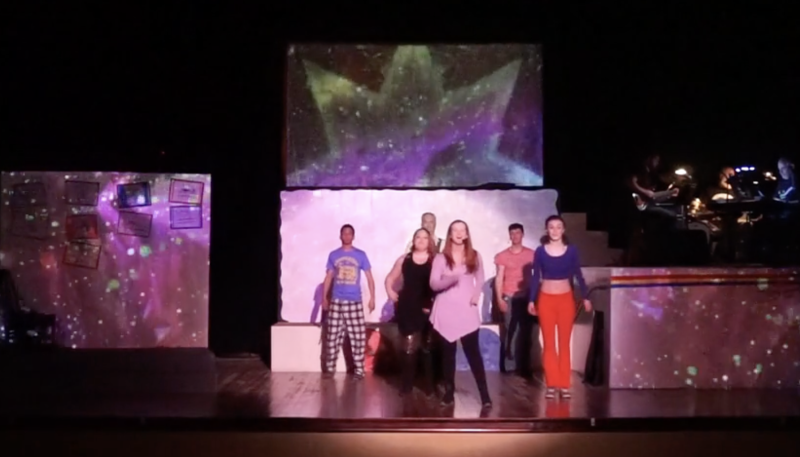 Performers on a stage with a large screen projecting a purple and green star, and a side screen with smaller images.