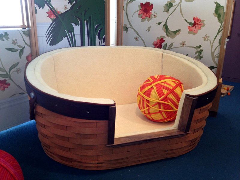 Woven basket pet bed with a cream fleece interior and a large red and yellow yarn ball inside.