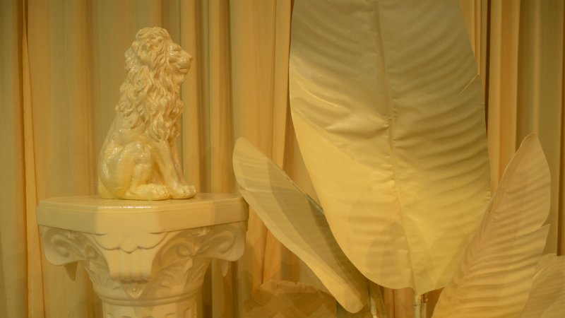 Monochromatic yellow artwork featuring a lion statue on a column and large fabric leaves against draped fabric.