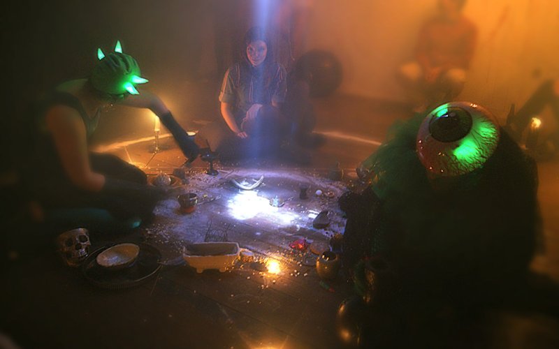 Three figures in a dimly lit room with ritualistic objects on the floor, including a glowing green eye and skull.