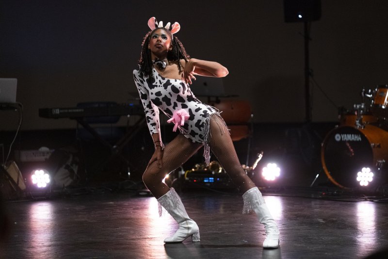 Performer in cow-print bodysuit, fishnets, and white boots poses on stage with musical instruments in the background.