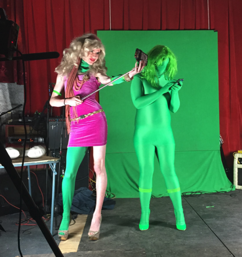 Two figures, one in a pink dress and green tights, the other fully in green, pose before a green screen and red curtain.