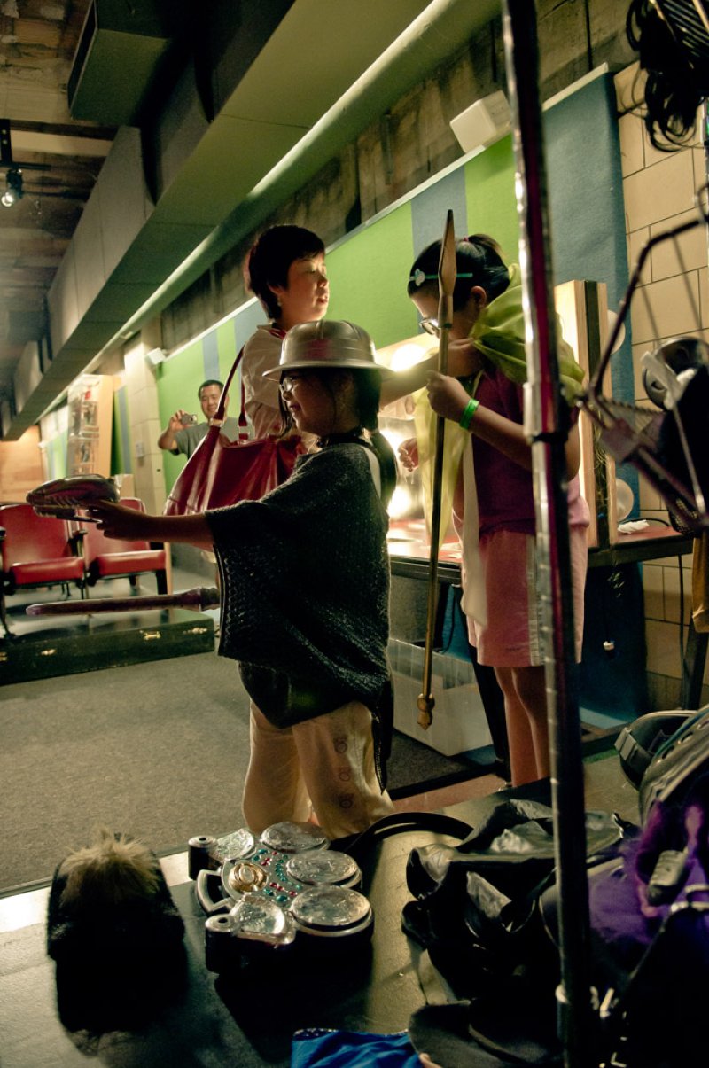 Three people in a dimly lit room, one wearing a silver helmet and holding a shoe, another in a pink skirt and green cape, and