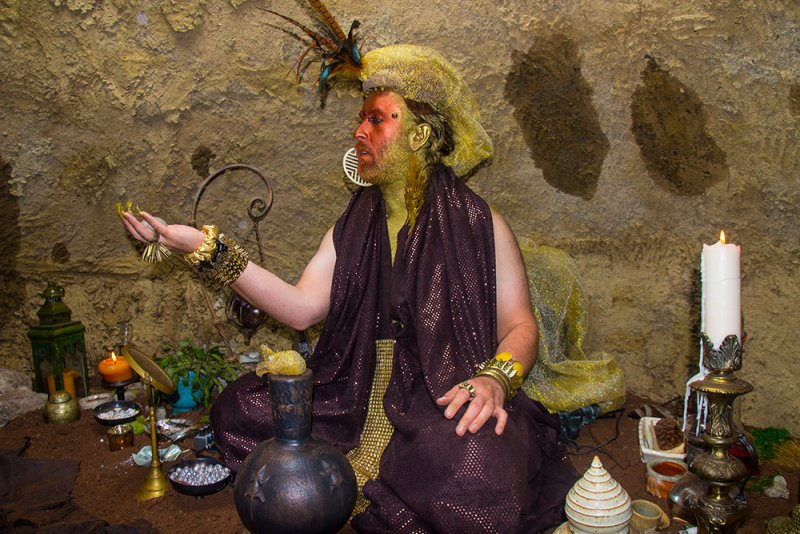 Performance artist in a cave, adorned with gold body paint, a feathered headdress, and a sequined robe, surrounded by rituali