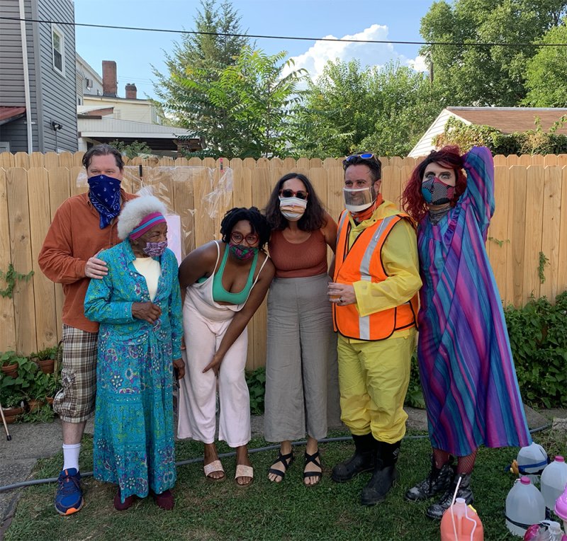 Six people in colorful, eclectic outfits and face masks pose in a grassy backyard with a wooden fence.