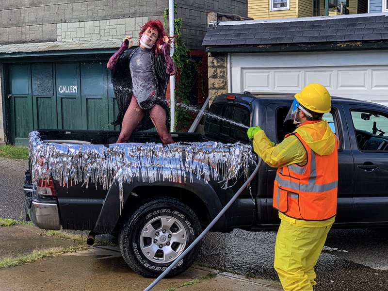 Person in drag with red hair and fishnets posing in a tinsel-decorated truck bed, being hosed by a worker in safety gear.