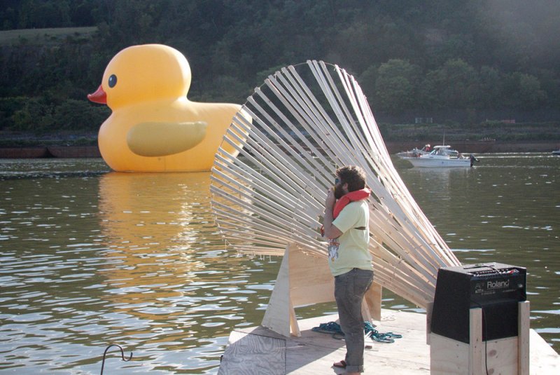 A man in a life vest stands on a raft next to a large wooden fan-like structure, facing a giant inflatable rubber duck on the