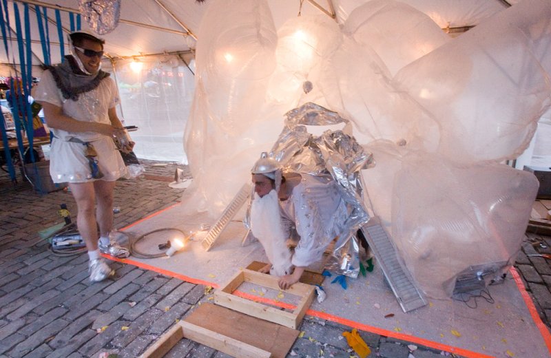 Two performers in costumes, one standing, one kneeling, surrounded by large white inflatable structures and scattered debris.