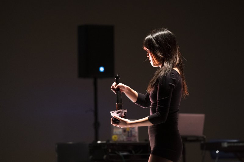 Woman in black dress holding a microphone over a cup, illuminated by a side light, with dark background and speaker.