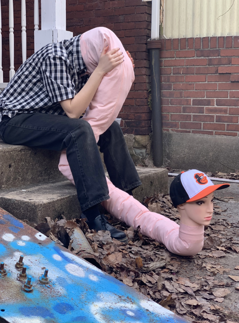 Person in plaid shirt and black jeans sitting on concrete steps, holding a pink fabric form that extends into a mannequin hea