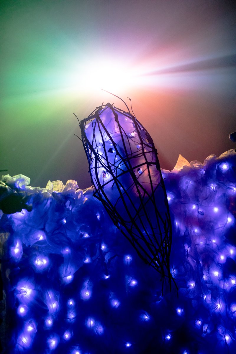 Sculpture of a dark, skeletal pod with purple light, surrounded by glowing blue lights and a green-to-red gradient background