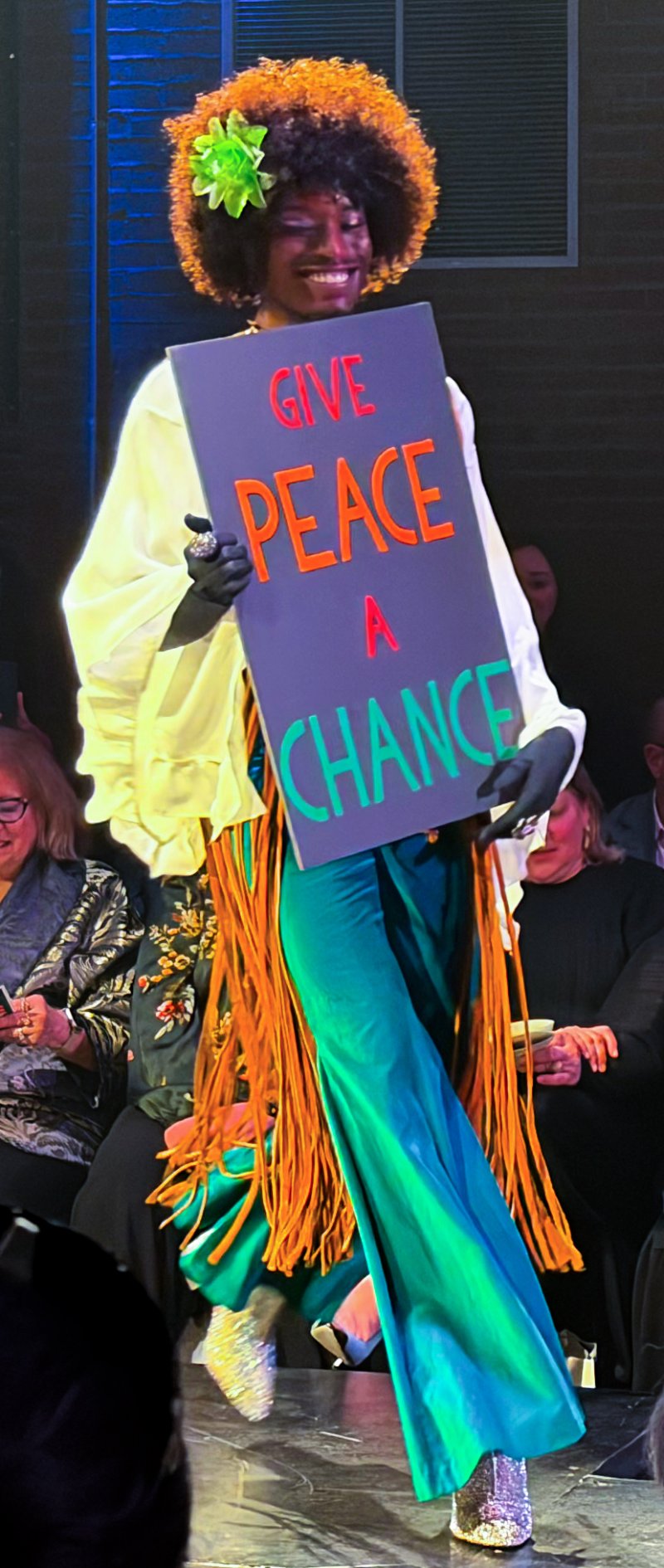 Smiling model with an orange afro, wearing a white blouse, green pants, and holding a "GIVE PEACE A CHANCE" sign.