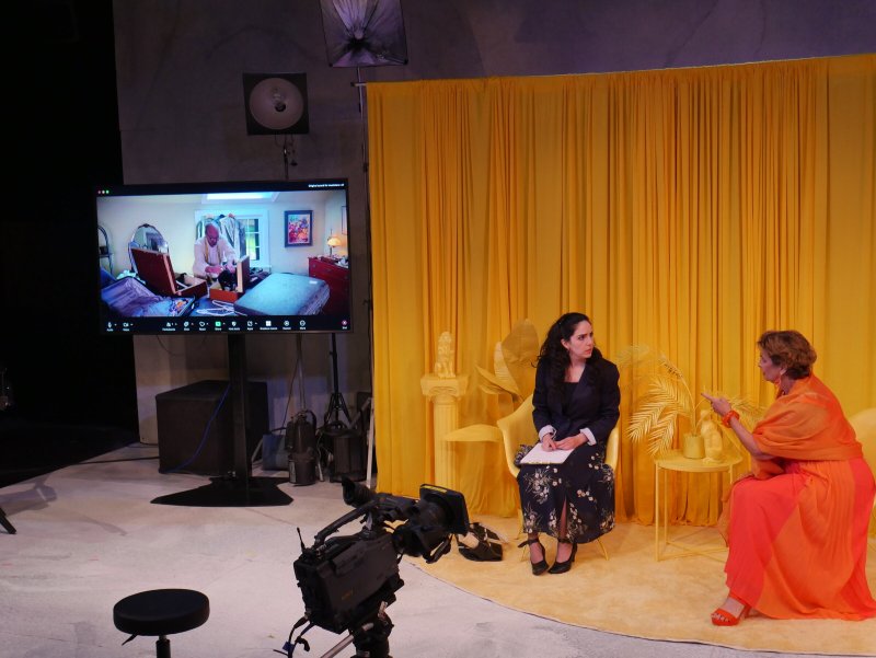 Two women in a yellow room, one in a navy suit, the other in an orange dress, conversing beside a large screen.