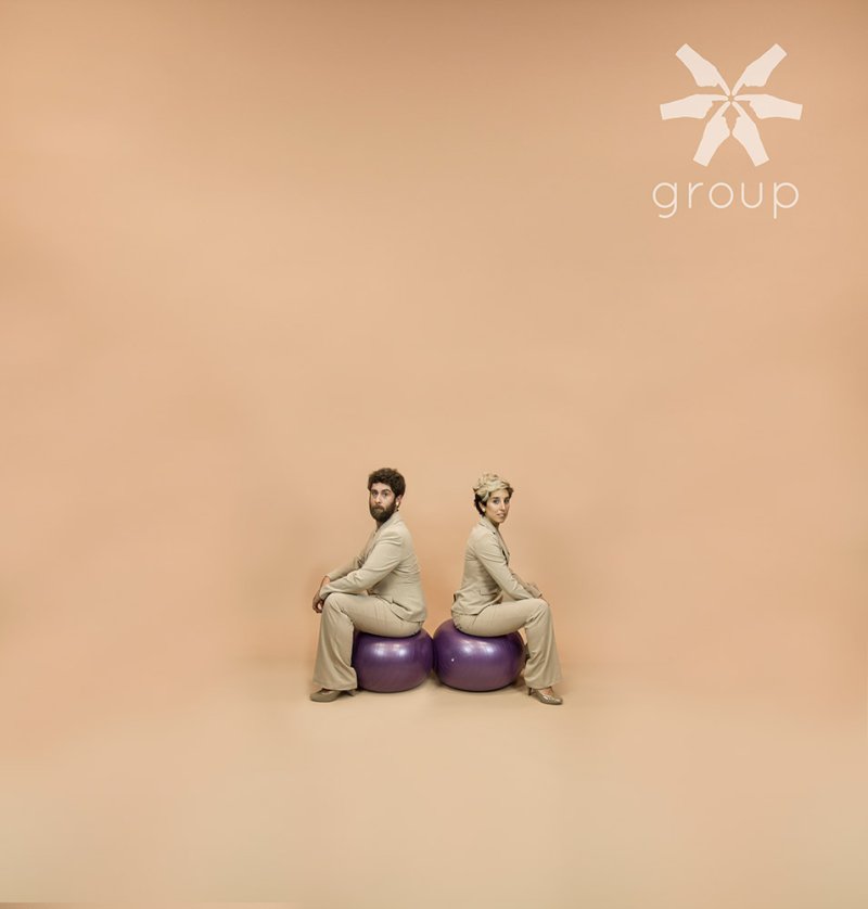 Two people in tan suits sit back-to-back on purple exercise balls against a peach background.