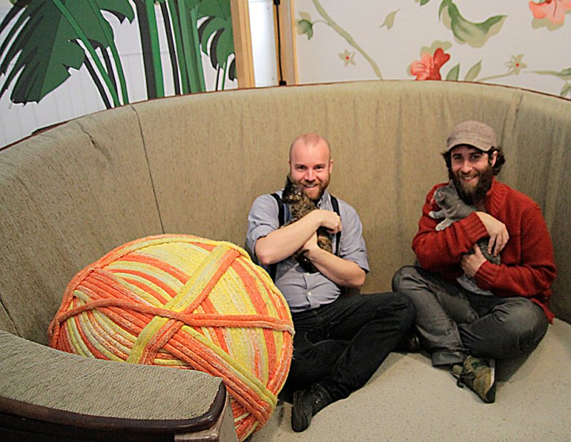 Two smiling men on a curved couch, each holding a kitten. A large, orange and yellow yarn ball sits beside them.