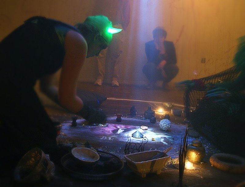 A person in a green glowing visor kneels over a ritualistic arrangement of objects, candles, and purple light on the floor.