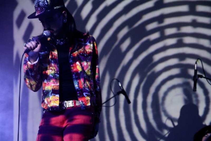Performer with a hat and colorful jacket sings into a microphone. Behind them is a projected concentric circle pattern. The performer wears red pants and a studded belt.