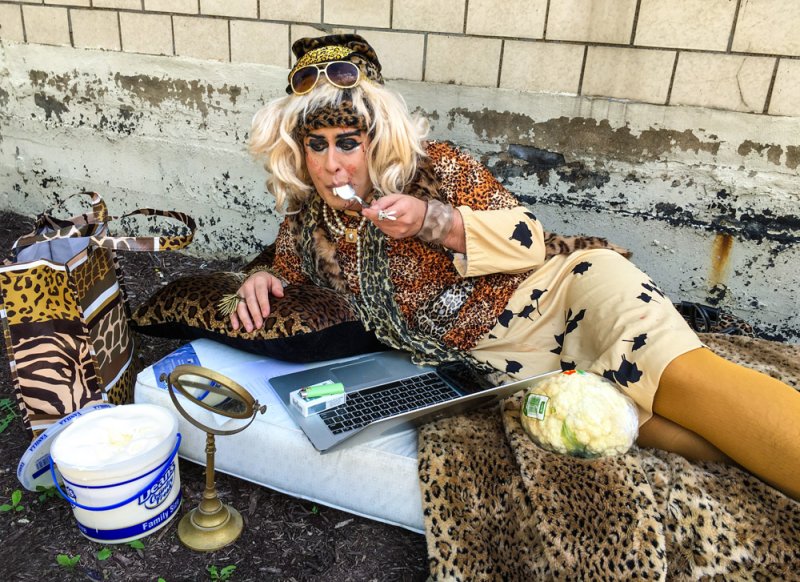 A person in drag with blonde hair, leopard print outfit, and sunglasses, reclines on a mattress outdoors, eating from a spoon