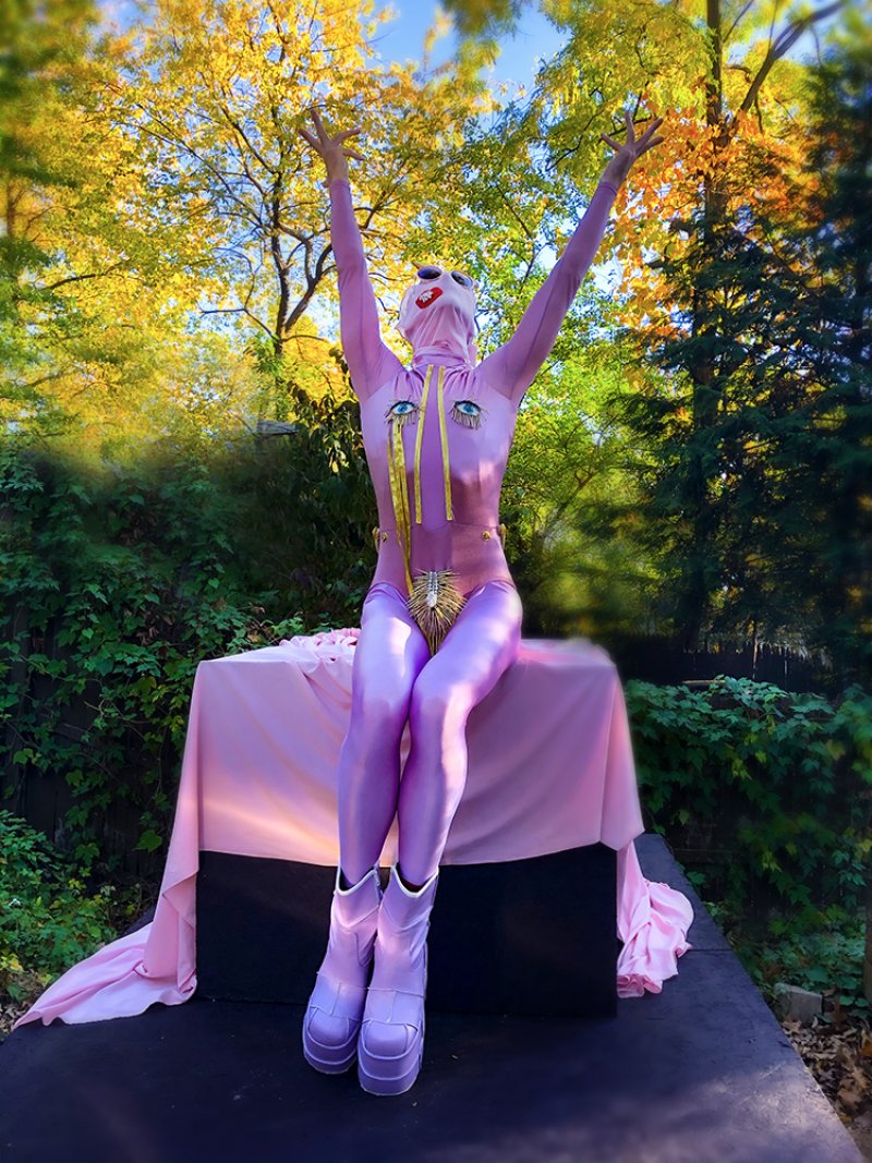 A figure in a pink and purple bodysuit with a white mask and gold embellishments sits on a draped pink box, arms raised, agai