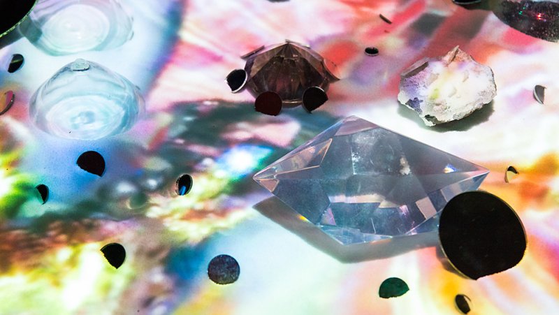 Abstract image of faceted clear and dark objects on a surface with colorful, blurred light reflections.