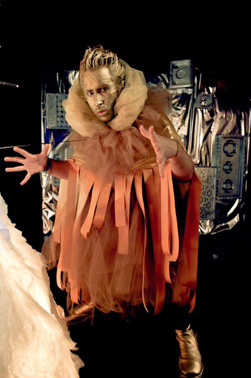 Performer in gold face paint and ruffled peach costume with flowing strips, against a metallic silver background.