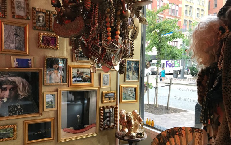 Eclectic wall display with numerous framed artworks, ornate golden frames, a beaded chandelier, and a storefront window.