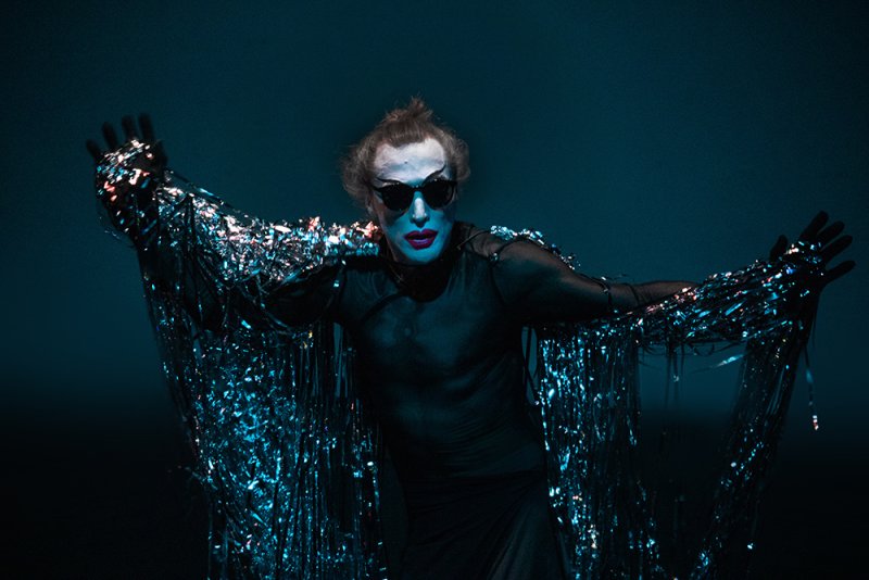 Performer in dark clothes, white face paint, red lips, and sunglasses, with arms outstretched, draped in shimmering silver ti