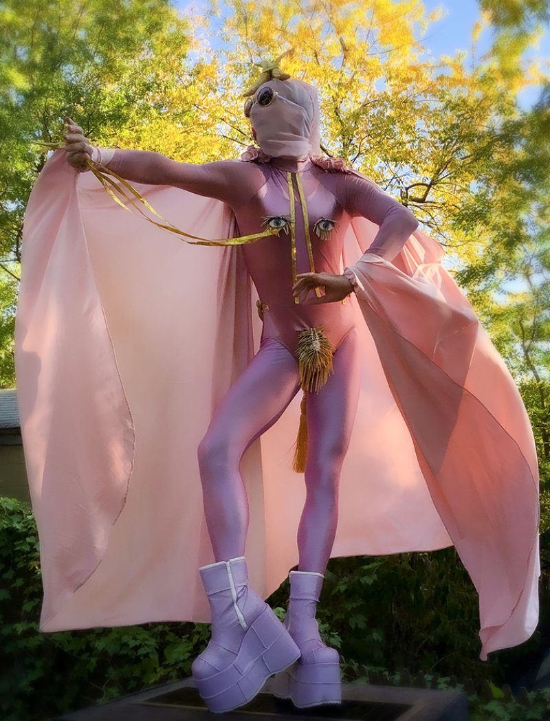 Person in pink bodysuit, cape, and lavender platform boots, with gold eyes and tassels on chest and groin, outdoors.