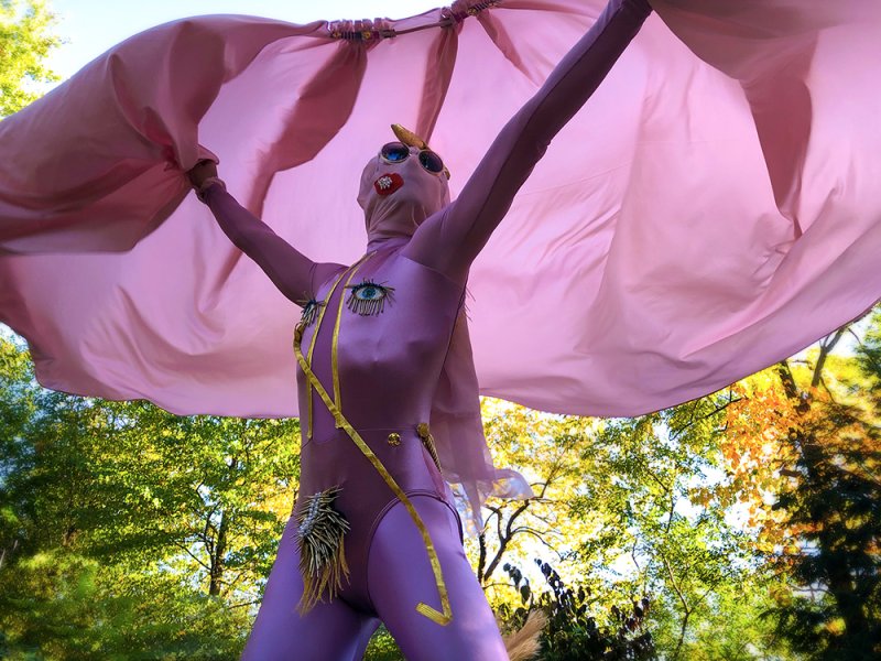 Performance artist in a pink bodysuit with golden accents, sunglasses, and a pink cape against a bright green and yellow tree
