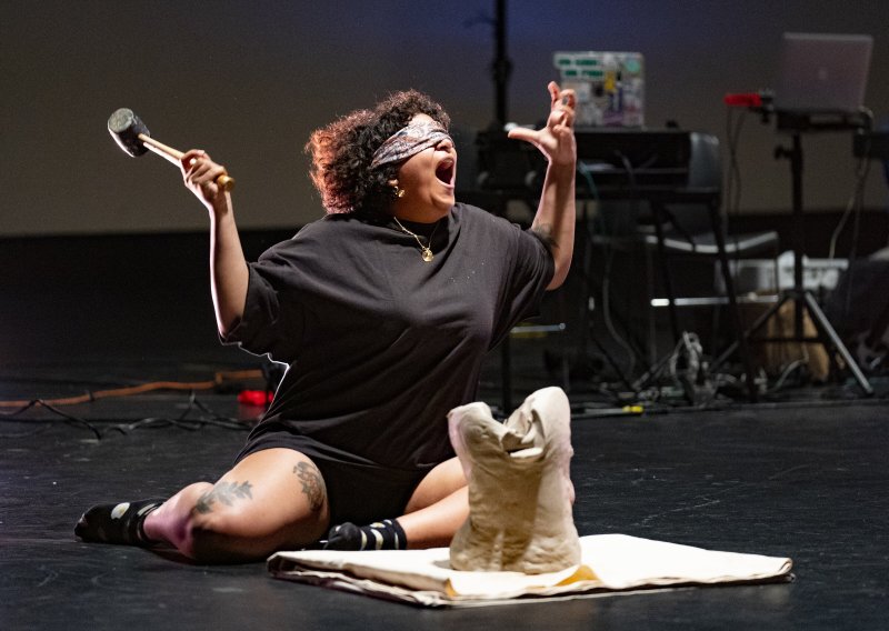 Performance art: Blindfolded figure with hammer, seated near sculpture on white cloth, dark stage.