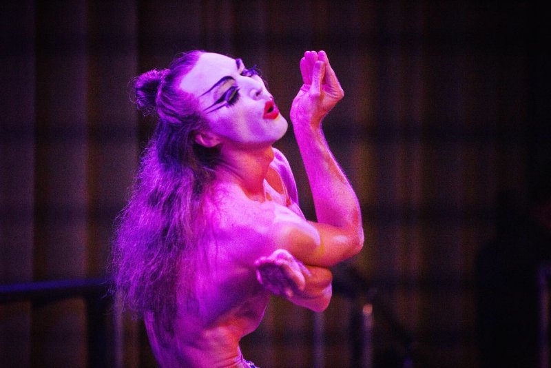 Performer in white face makeup, dramatic black eyeliner, and red lipstick, illuminated by purple light.