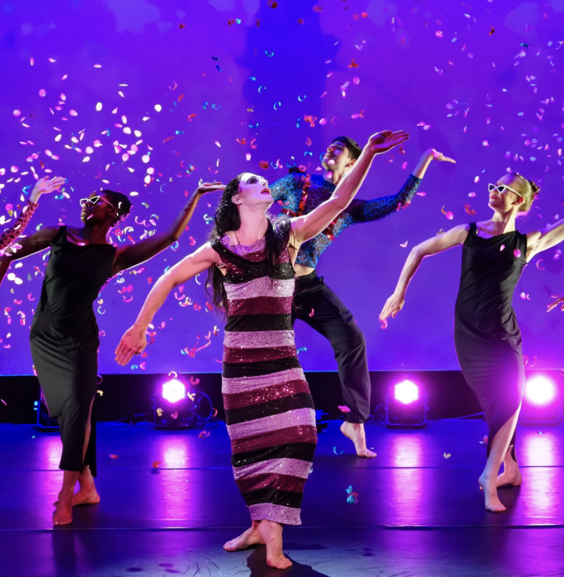 Performance art piece with four dancers on stage under falling confetti. Three wear dark dresses, one a patterned shirt and pants; the central dancer has a horizontally striped dress. The backdrop is a bright blue/purple.