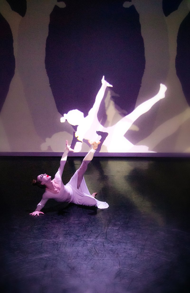 A dancer in a white dress on a dark stage, illuminated by a purple light, with a projected silhouette of another dancer on th
