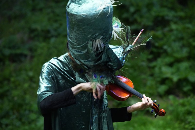 Person in a shimmering green costume with a tall hat and peacock feathers playing a violin against a blurred green background