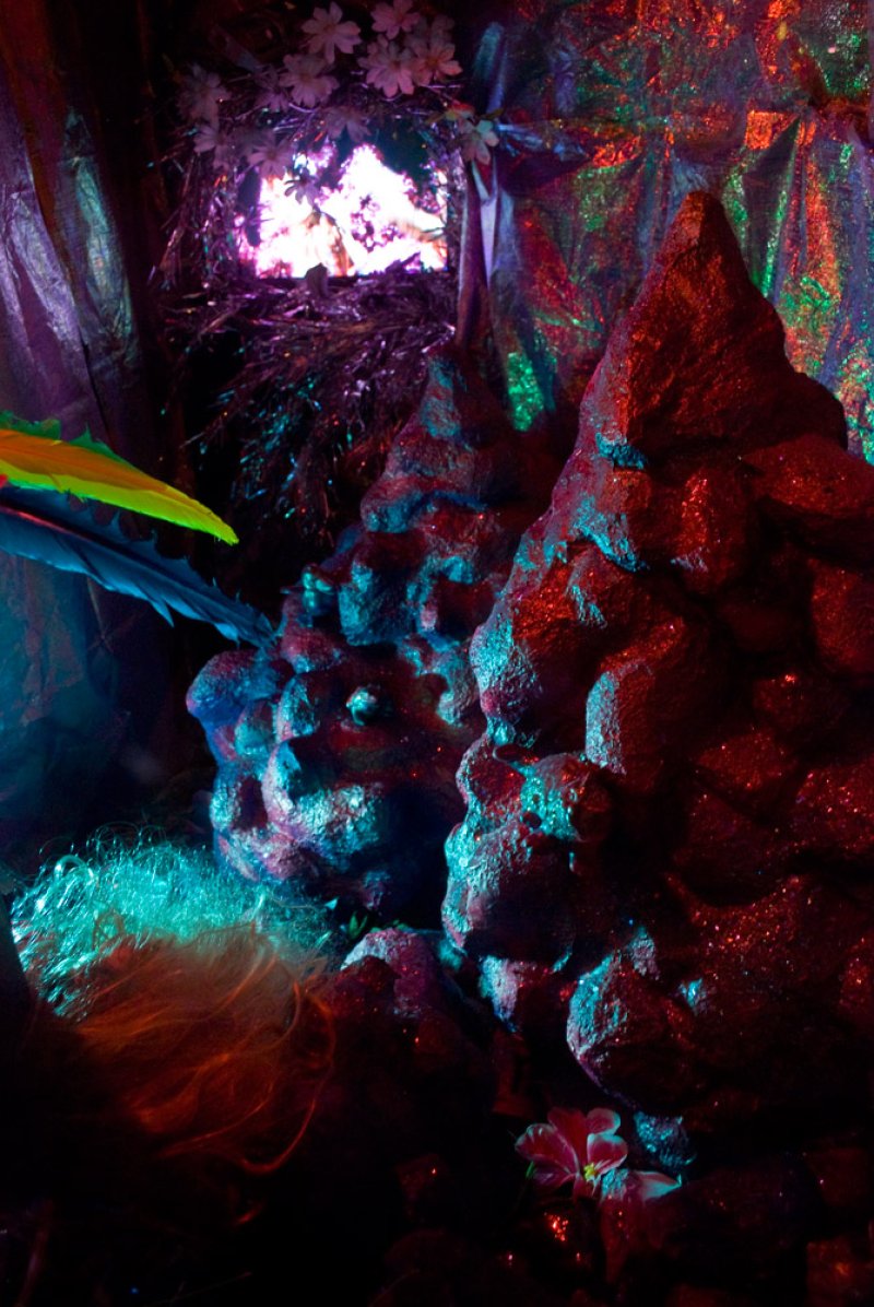 Sculptural installation with two red, rock-like forms, illuminated by blue and red light, against a dark, shimmering backgrou