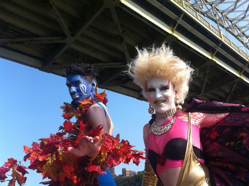 Two performers under a bridge, one in blue body paint and red leaves, the other in drag with a blonde wig and pink top.