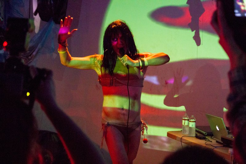 Performer in a dark wig and minimal clothing, bathed in green and red stage lights, singing into a microphone.