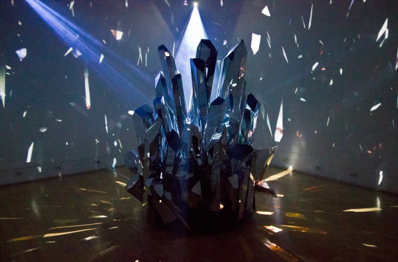 Sculptural installation of dark, faceted crystal forms illuminated by spotlights, casting reflections and light shards on wal