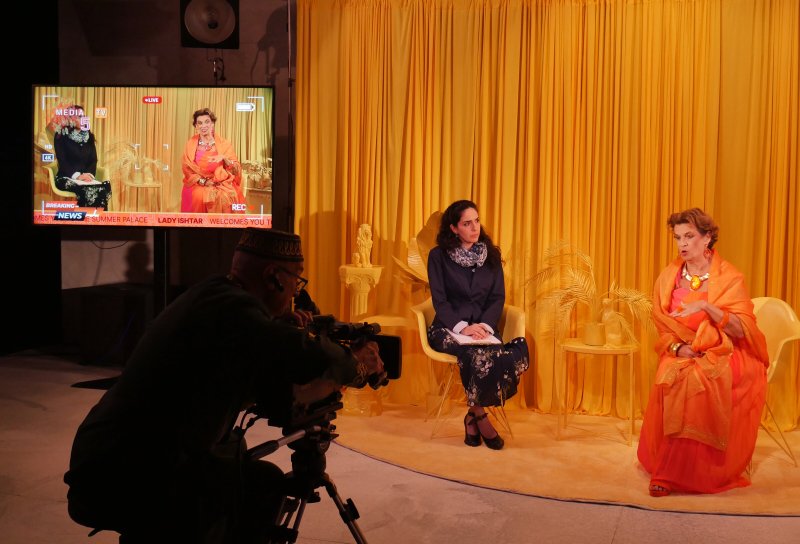 Installation view: Two women seated on yellow chairs against a yellow curtain backdrop, one in orange attire. A cameraman films them. A monitor displays the same scene with news graphics.