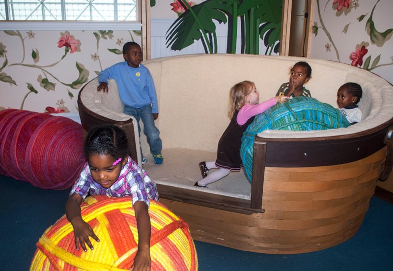 Children playing with large yarn balls in a boat-shaped couch and on the floor, against a floral wallpaper backdrop.