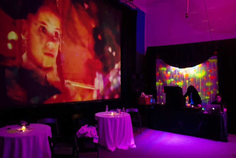An art installation with a large projection of a man's face in red and orange hues, two round tables with white tablecloths, 